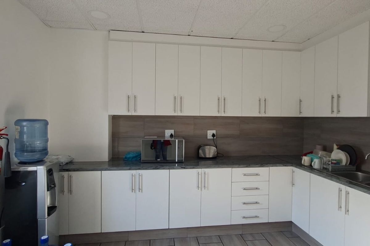 A clean and functional office kitchenette featuring white cabinetry, laminate countertops, a stainless steel double sink, and a tiled backsplash.
