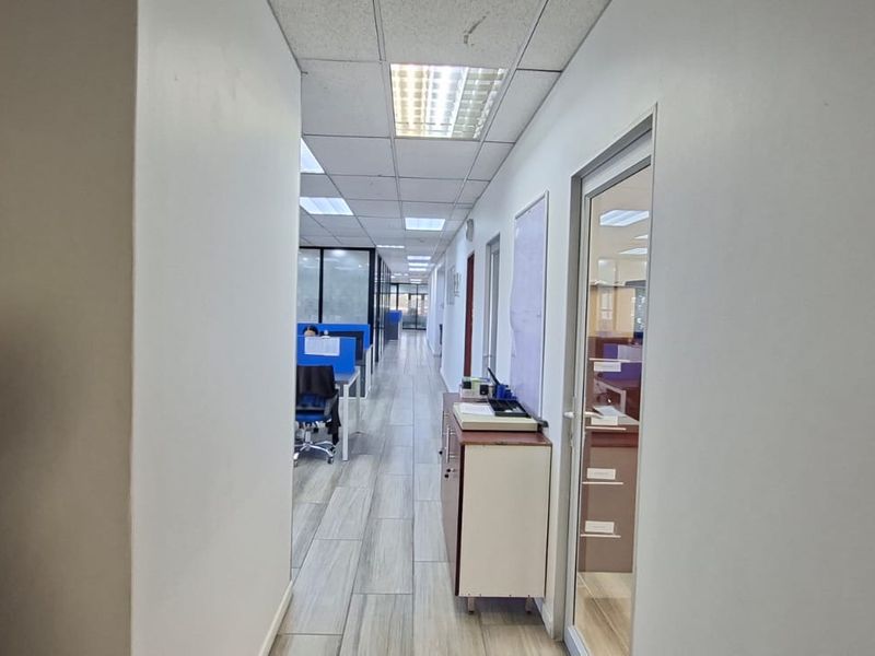 A functional office hallway featuring tiled flooring, suspended ceiling with recessed lighting, and glass partition doors leading into workspace areas.