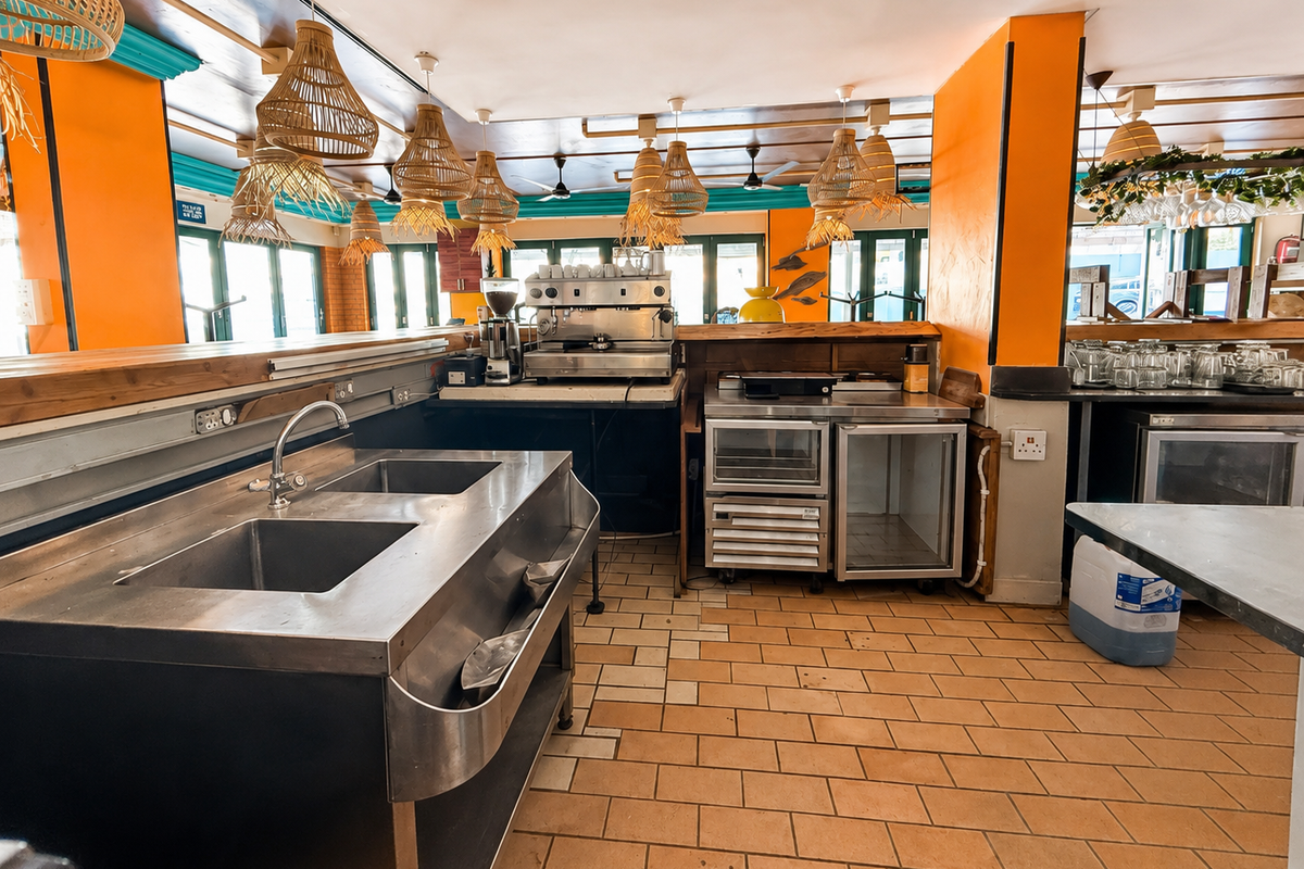 A functional restaurant kitchen area featuring stainless steel sinks, commercial coffee equipment, under-counter refrigeration, and tiled flooring with decorative pendant lighting.