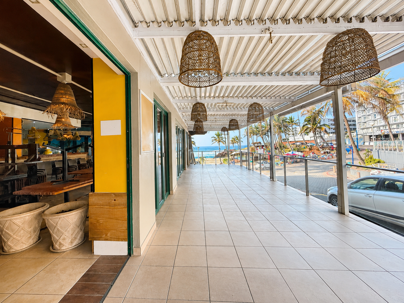 A spacious covered outdoor patio area featuring tiled flooring, a corrugated metal ceiling, and a glass balustrade with views of the beach.