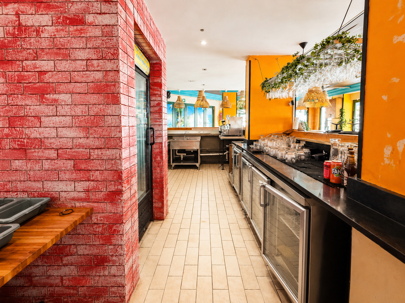 A functional restaurant bar area featuring a stainless steel prep sink, under-counter refrigeration units, and a brick feature wall with overhead glassware storage.