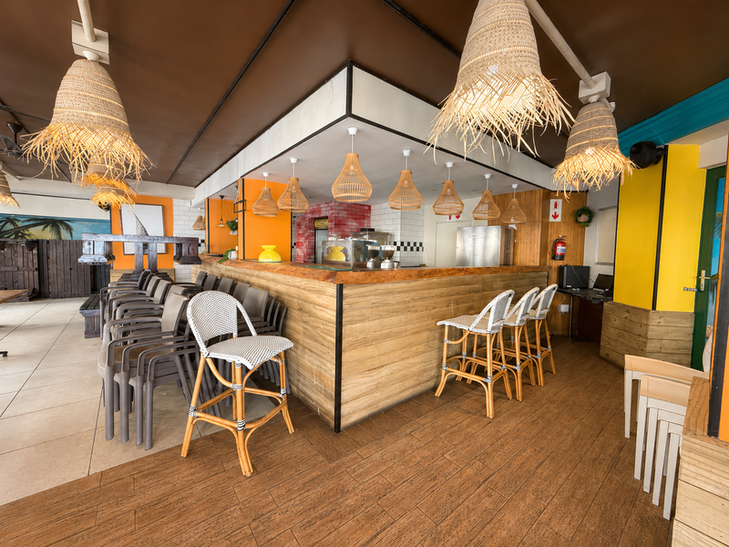 A vibrant restaurant interior featuring a prominent wooden bar counter, mixed flooring, and decorative pendant lighting, creating a casual dining atmosphere.