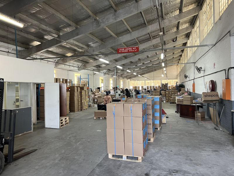 Spacious industrial warehouse featuring high ceilings with exposed steel trusses, concrete flooring, and functional partitioned office space, suitable for storage or manufacturing operations.