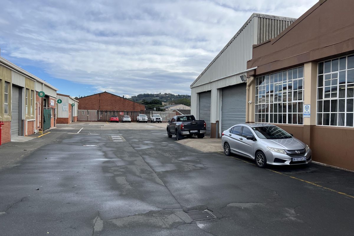 Industrial yard area featuring multiple roller shutter doors, steel-framed windows, and paved access space between warehouse buildings.