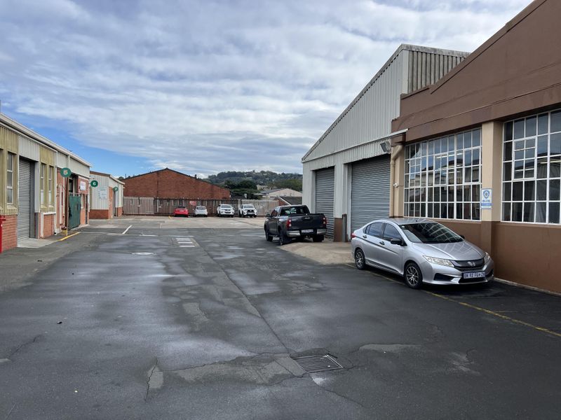 Industrial yard area featuring multiple roller shutter doors, steel-framed windows, and paved access space between warehouse buildings.