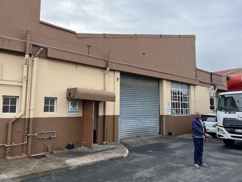 Industrial warehouse exterior featuring a large roller shutter door, steel-framed windows, and an asphalt driveway access point.