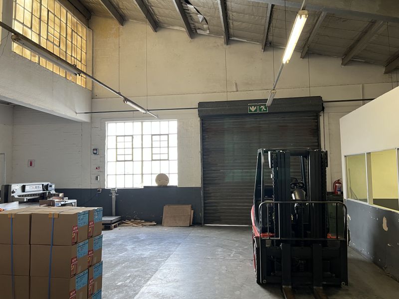 Spacious industrial warehouse area featuring high ceilings with exposed steel trusses, concrete flooring, large windows, and a secure roller shutter door for loading access.
