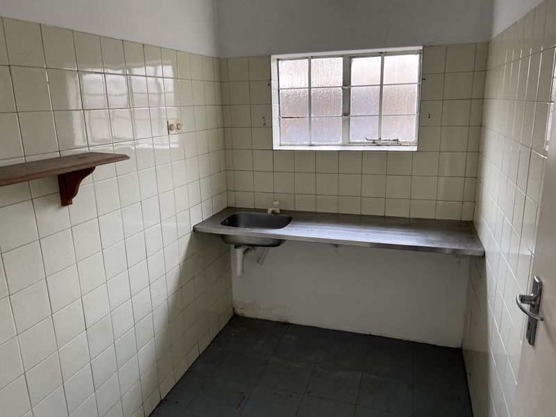 A compact, empty scullery or utility area featuring a stainless steel sink, tiled walls, and a metal-framed window.