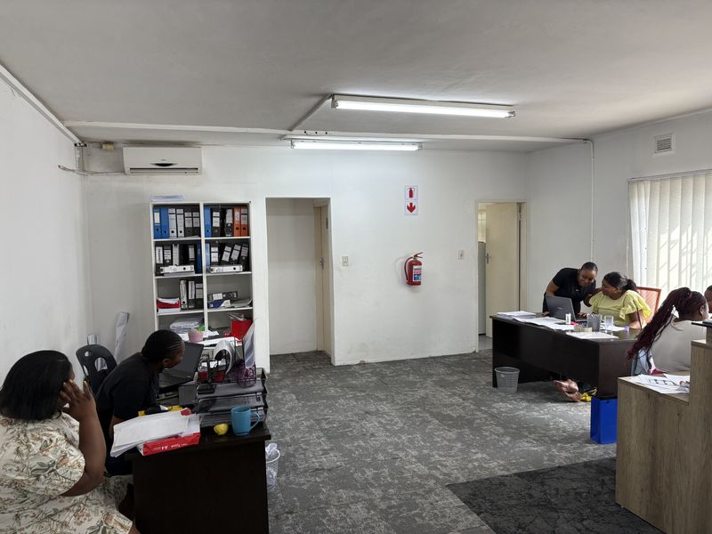 A functional office space featuring carpeted flooring, air conditioning, and overhead fluorescent lighting, currently configured with multiple workstations.