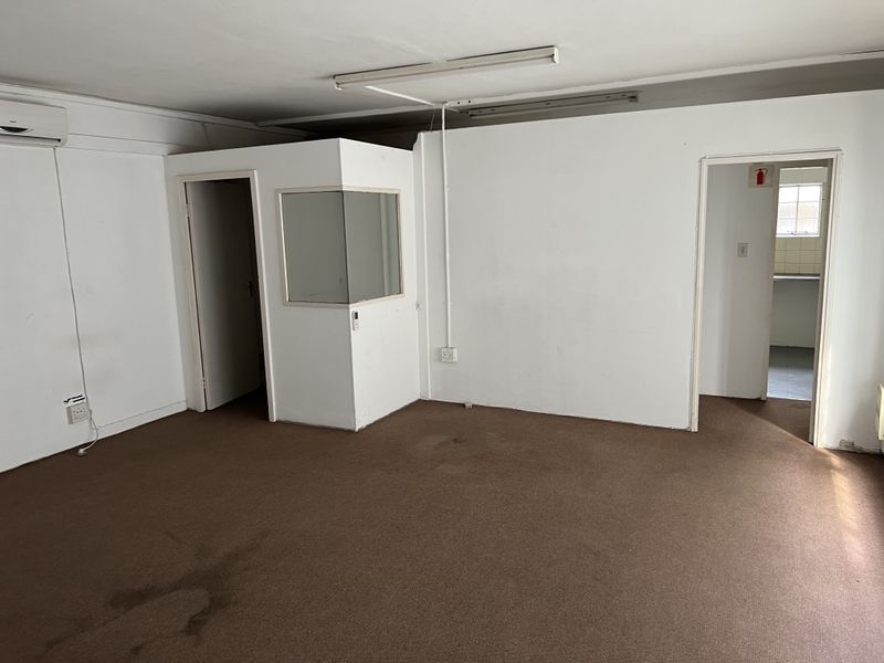 An empty office space featuring carpet flooring, a partitioned cubicle with an internal window, and wall-mounted air conditioning.