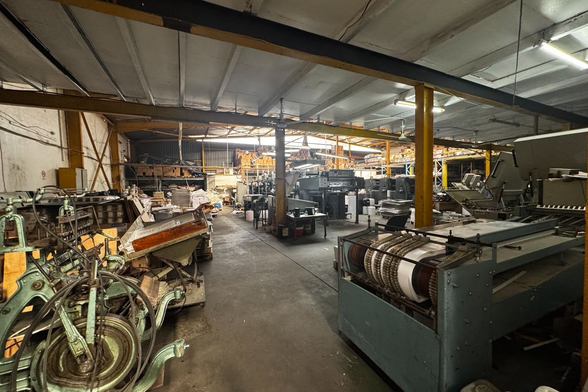 Spacious industrial warehouse area featuring exposed steel structural beams, concrete flooring, and overhead fluorescent lighting, currently utilized for manufacturing or printing operations.