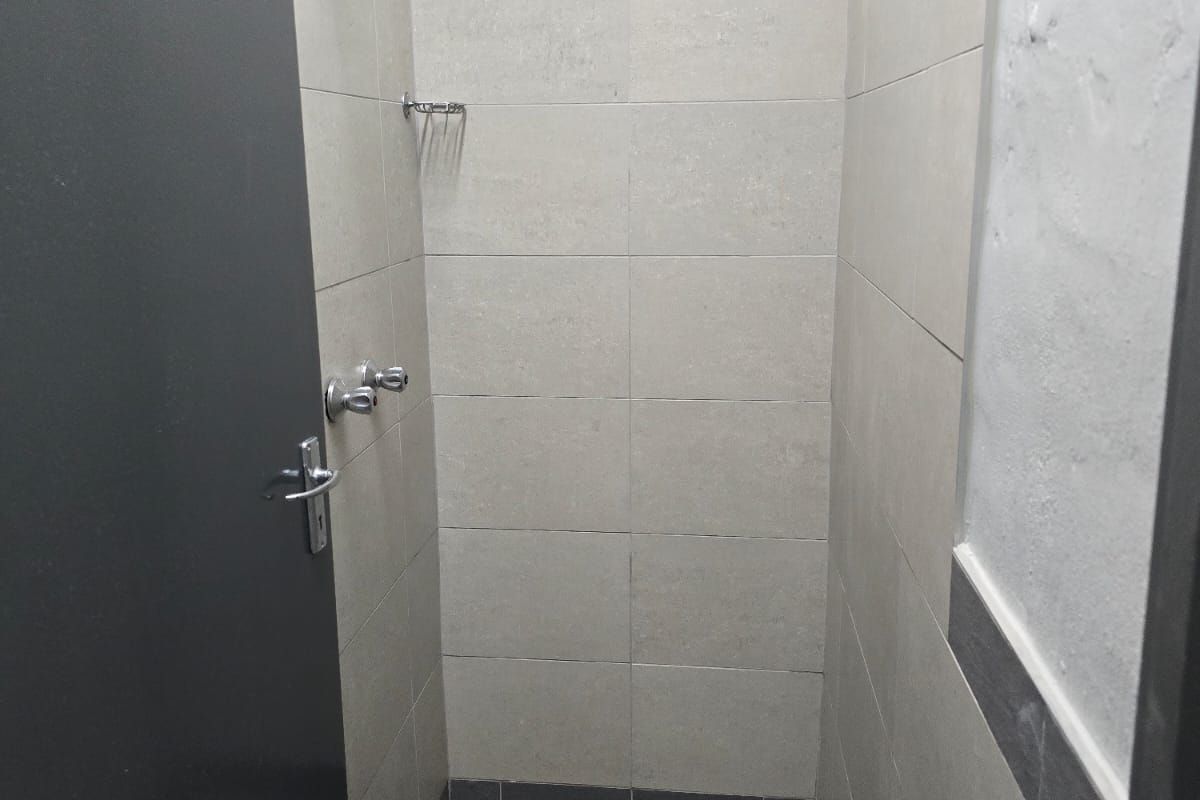 A functional industrial-style shower stall featuring neutral wall tiling, a floor drain, and basic chrome shower fixtures.