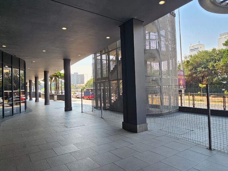 A modern covered retail walkway featuring tiled flooring, recessed lighting, and a glass storefront facade with an adjacent metal mesh stairwell structure.