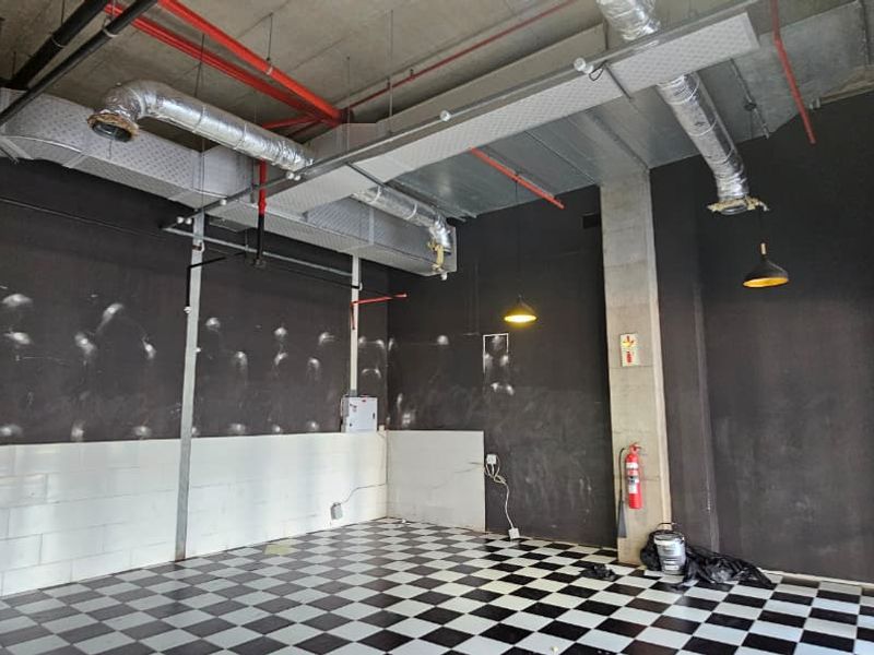 An empty, industrial-style retail space featuring exposed ductwork, checkerboard tiled flooring, and dark painted walls, suitable for a variety of commercial fit-outs.