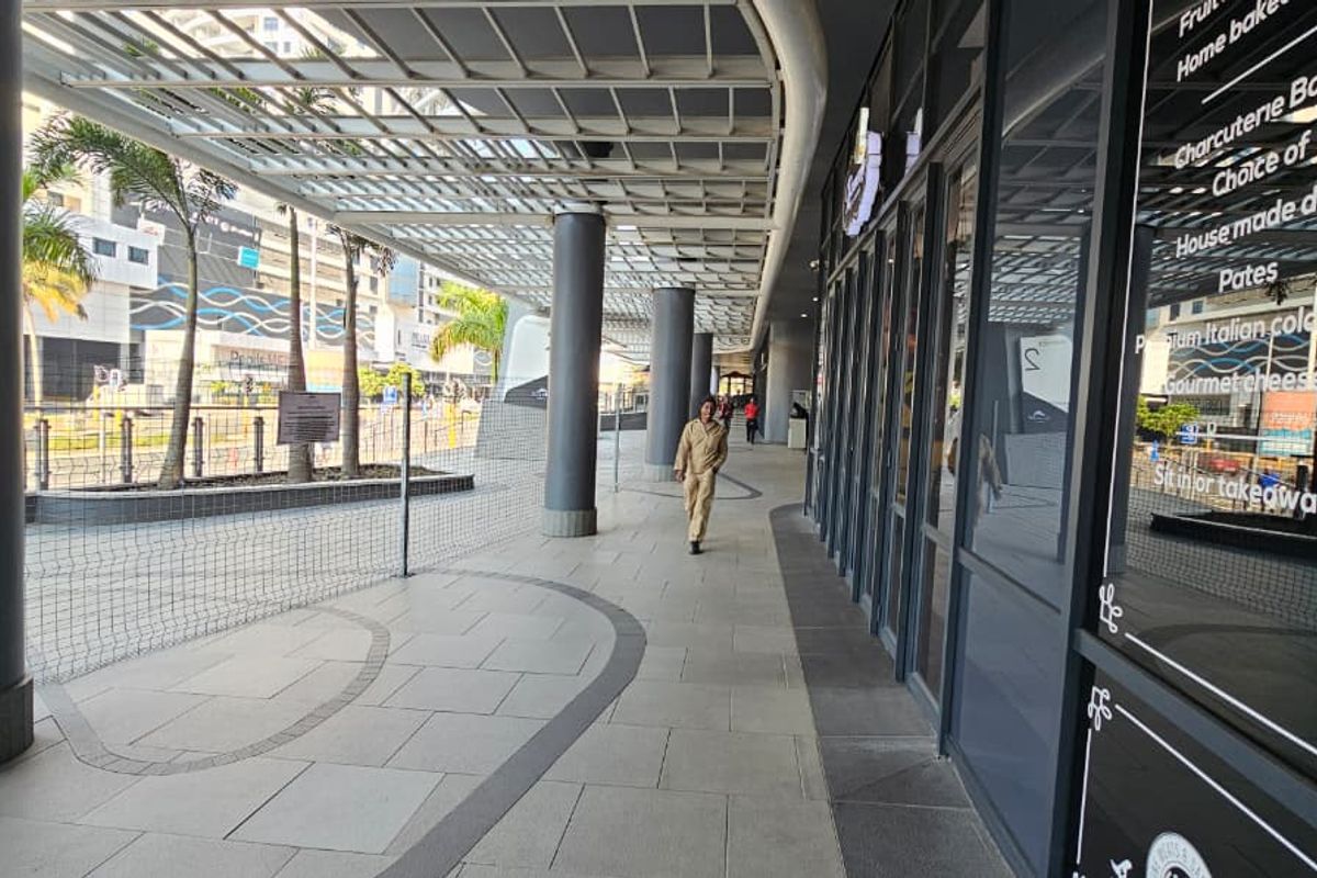A covered outdoor walkway and patio area featuring modern tiled flooring, structural metal columns, and a glass storefront facade for a retail space.