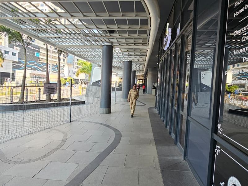 A covered outdoor walkway and patio area featuring modern tiled flooring, structural metal columns, and a glass storefront facade for a retail space.