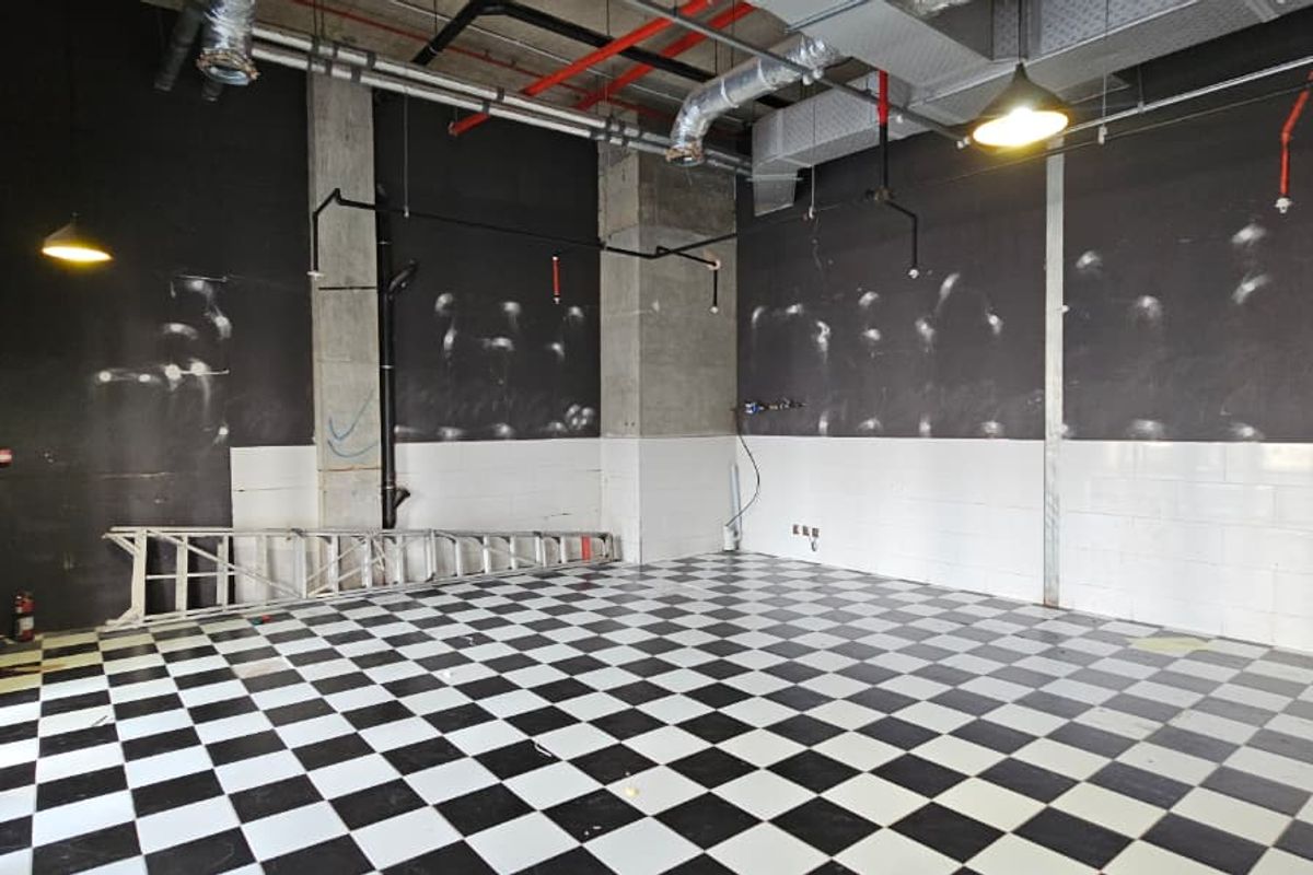An empty retail space featuring an industrial-style exposed ceiling, checkered tile flooring, and concrete structural columns, suitable for a variety of commercial uses.