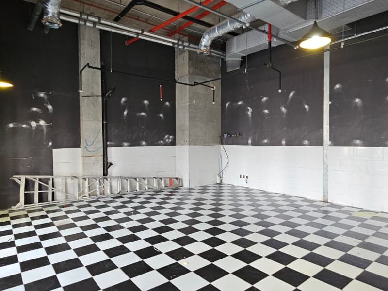An empty retail space featuring an industrial-style exposed ceiling, checkered tile flooring, and concrete structural columns, suitable for a variety of commercial uses.