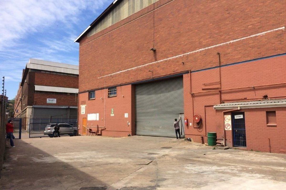 Rear exterior view of an industrial warehouse featuring a large roller shutter door, brick facade, and concrete yard area.