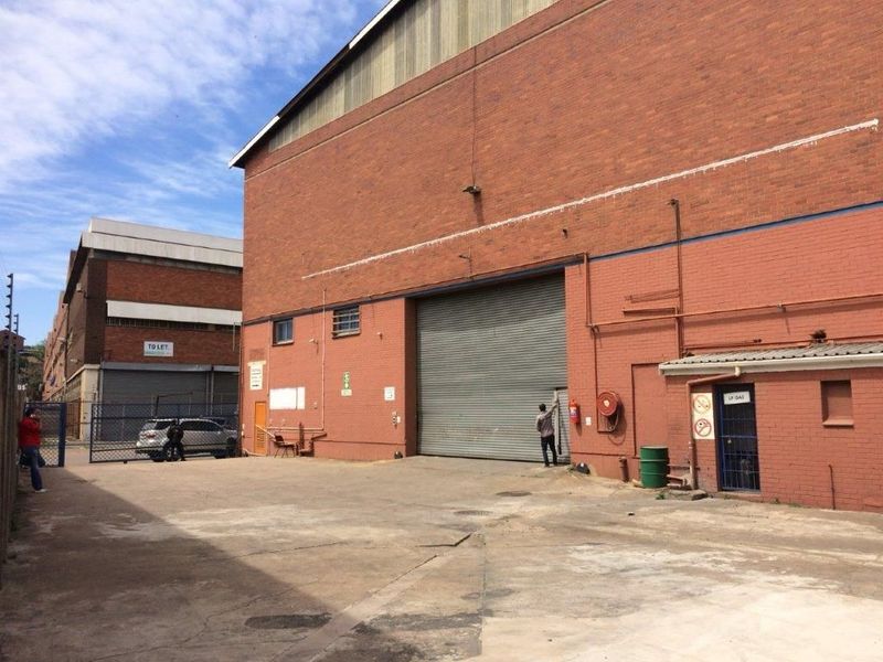 Rear exterior view of an industrial warehouse featuring a large roller shutter door, brick facade, and concrete yard area.