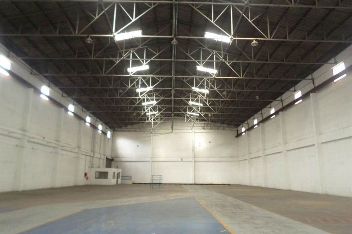 Spacious industrial warehouse featuring high ceilings with exposed steel trusses, high-bay lighting, concrete flooring, and a small partitioned office area.