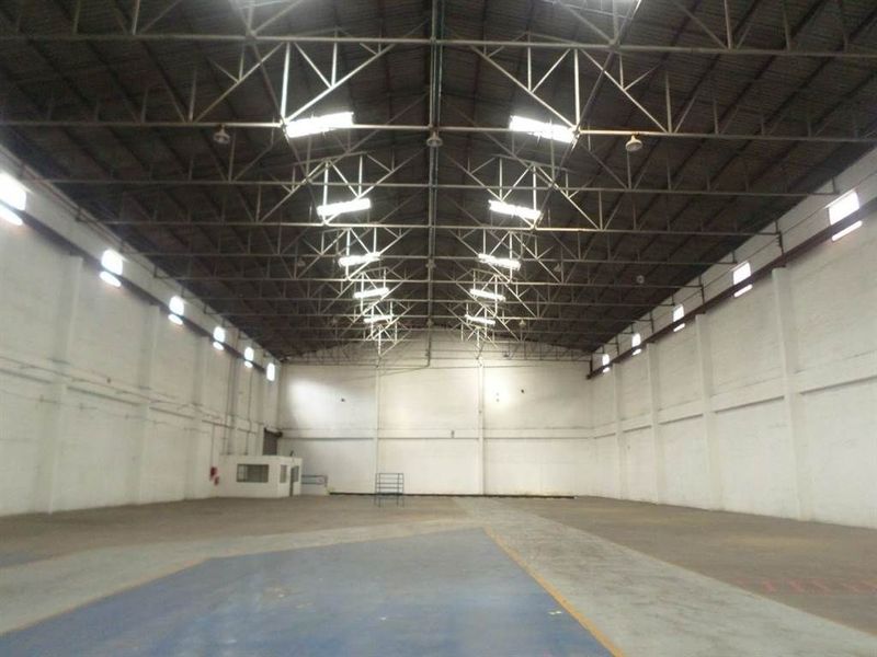 Spacious industrial warehouse featuring high ceilings with exposed steel trusses, high-bay lighting, concrete flooring, and a small partitioned office area.