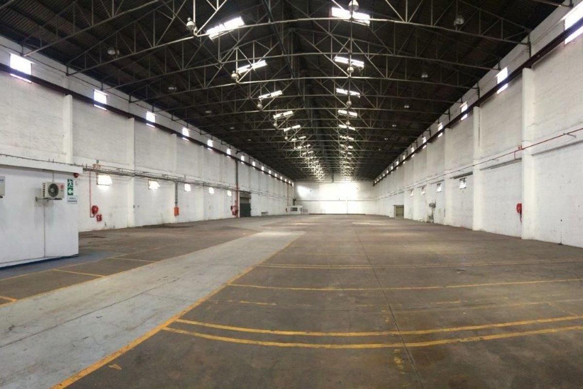 Large open-plan industrial warehouse space featuring high exposed steel truss ceilings, durable concrete flooring, and wall-mounted climate control units.