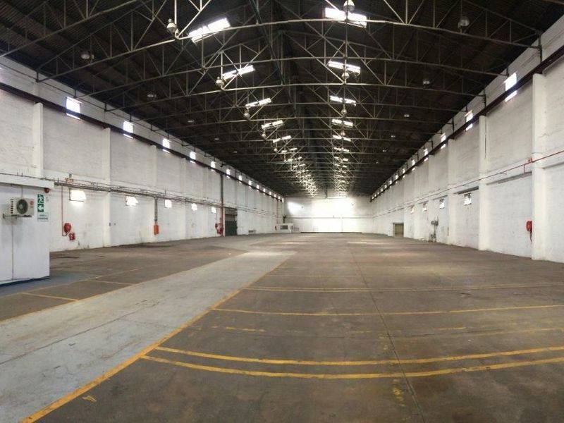 Large open-plan industrial warehouse space featuring high exposed steel truss ceilings, durable concrete flooring, and wall-mounted climate control units.