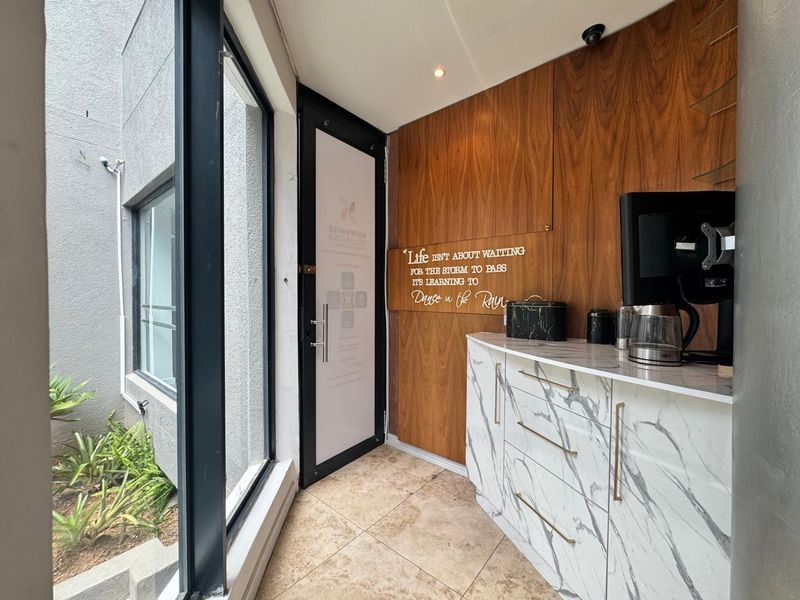 Modern office kitchenette area featuring marble-look cabinetry, wood veneer wall paneling, and large windows overlooking a small garden space.