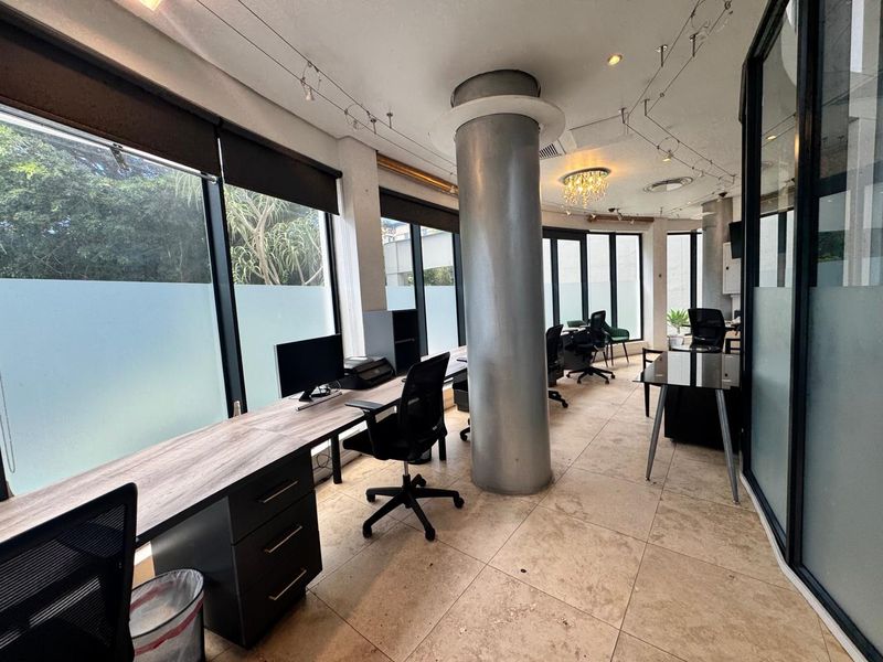 Modern open-plan office space featuring large windows, structural columns, tiled flooring, and glass partitions, offering a bright and professional work environment.