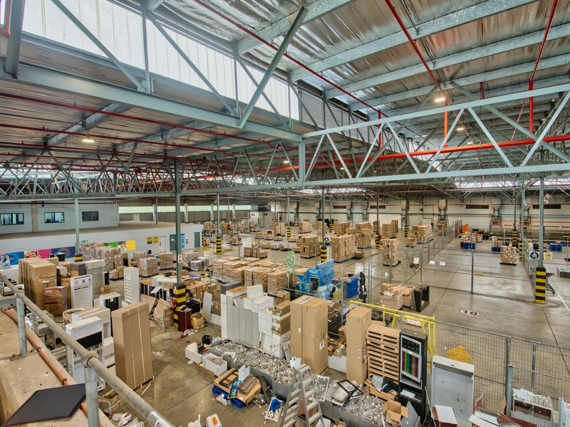 Large industrial warehouse space featuring high ceilings with exposed steel trusses, fire sprinkler systems, skylights, and concrete flooring with partitioned storage areas.