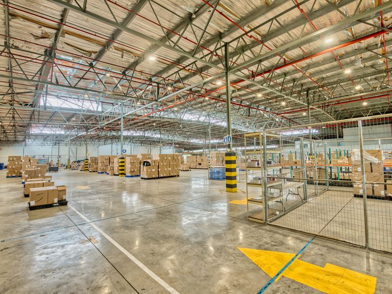 Large industrial warehouse space featuring high exposed steel truss ceilings, fire sprinkler systems, polished concrete floors, and secure metal mesh partitioning.