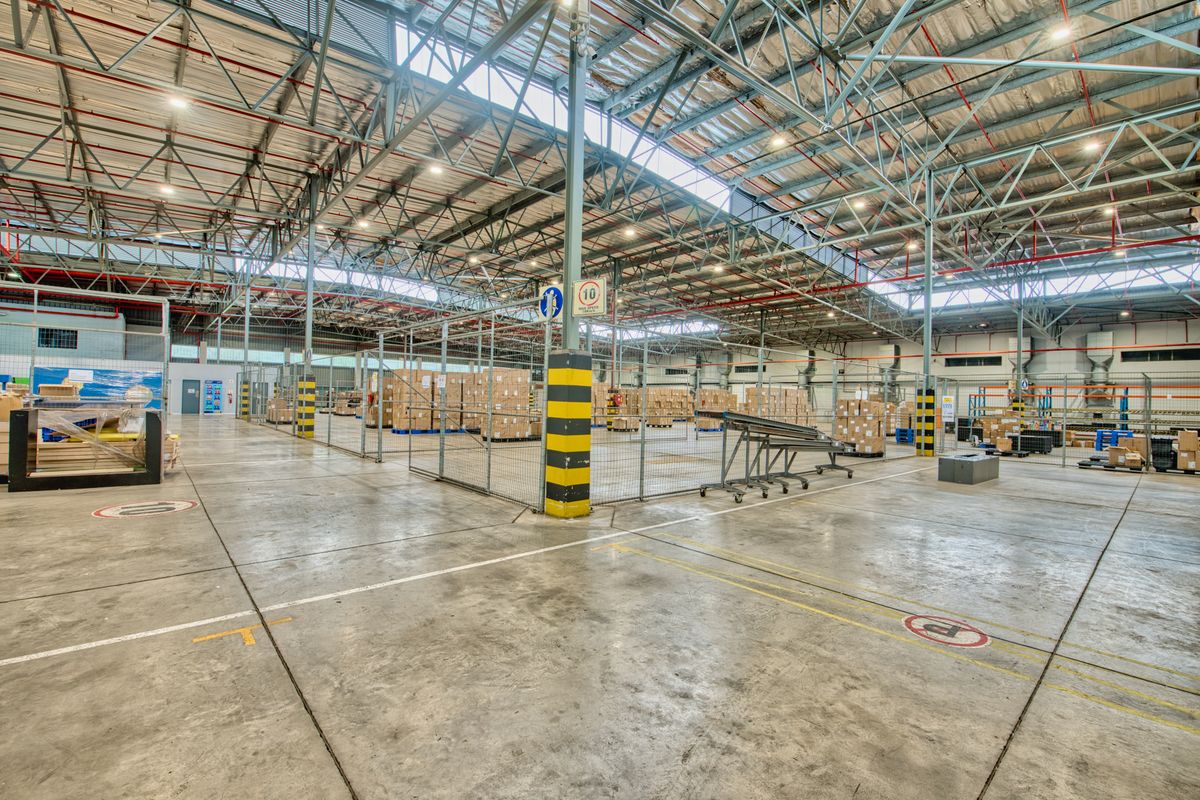 Spacious industrial warehouse facility featuring high exposed steel truss ceilings, durable concrete flooring, fire sprinkler systems, and secure wire mesh partitioned storage areas.