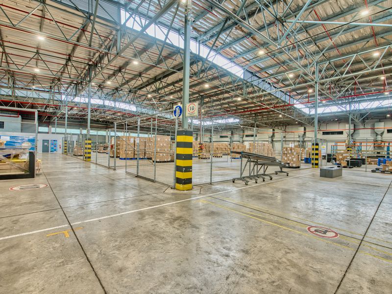 Spacious industrial warehouse facility featuring high exposed steel truss ceilings, durable concrete flooring, fire sprinkler systems, and secure wire mesh partitioned storage areas.