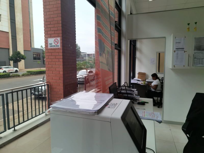 A commercial office space featuring large glass windows, exposed brick structural elements, and tiled flooring, providing a bright and functional work environment.