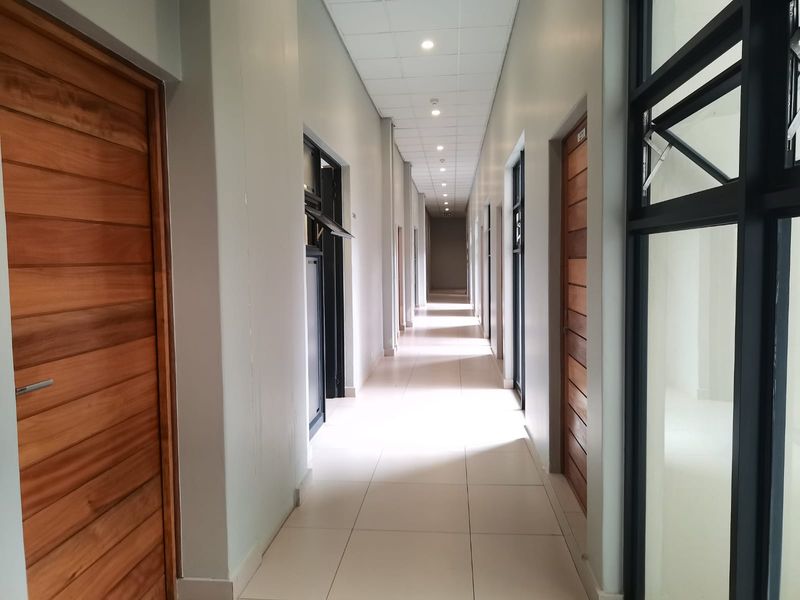 A clean, modern commercial hallway featuring tiled flooring, recessed downlights, and multiple timber doors leading to individual office or utility spaces.
