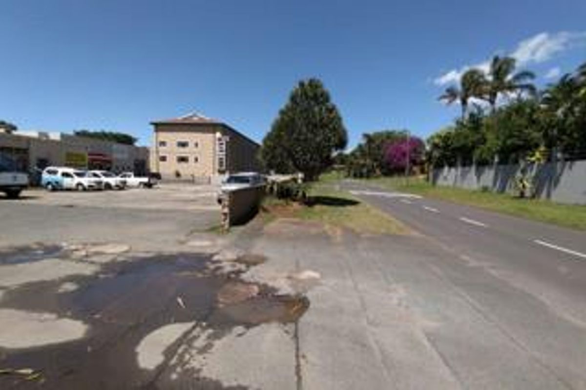 Street view of the retail property showing the asphalt parking area, boundary wall, and adjacent road access.