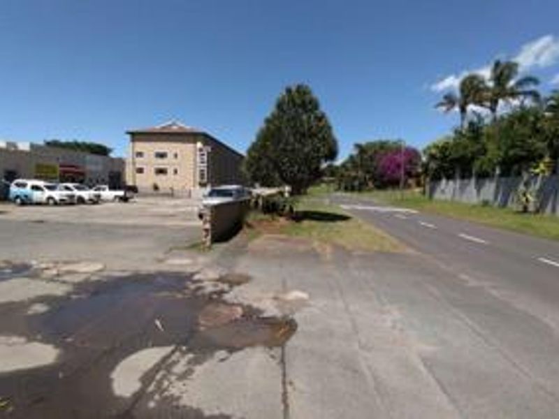 Street view of the retail property showing the asphalt parking area, boundary wall, and adjacent road access.