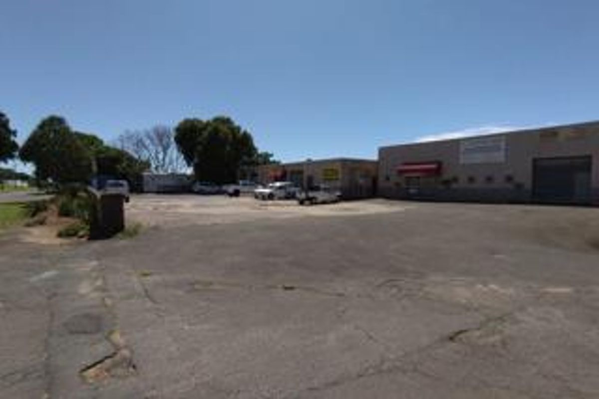 Exterior view of a commercial retail property featuring a large asphalt parking area and multiple access points with roller shutter doors.