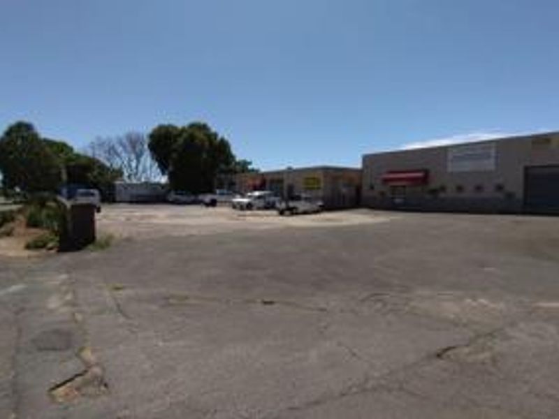 Exterior view of a commercial retail property featuring a large asphalt parking area and multiple access points with roller shutter doors.