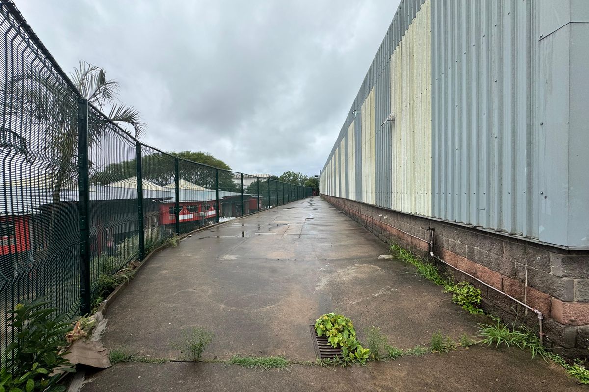 A narrow exterior side passage of an industrial building featuring corrugated metal walls, concrete block foundation, and security fencing.