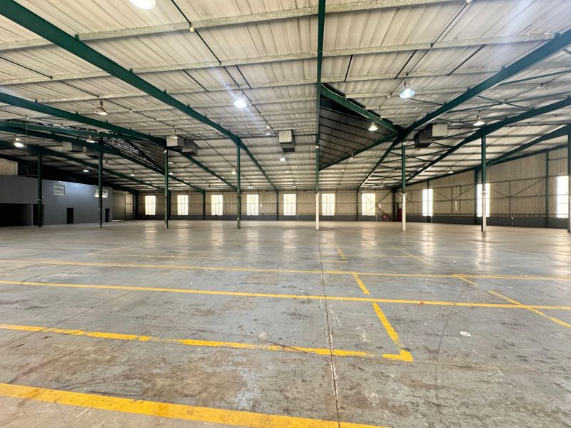 Expansive industrial warehouse space featuring high ceilings with exposed steel trusses, concrete flooring with marked bays, and functional overhead lighting.
