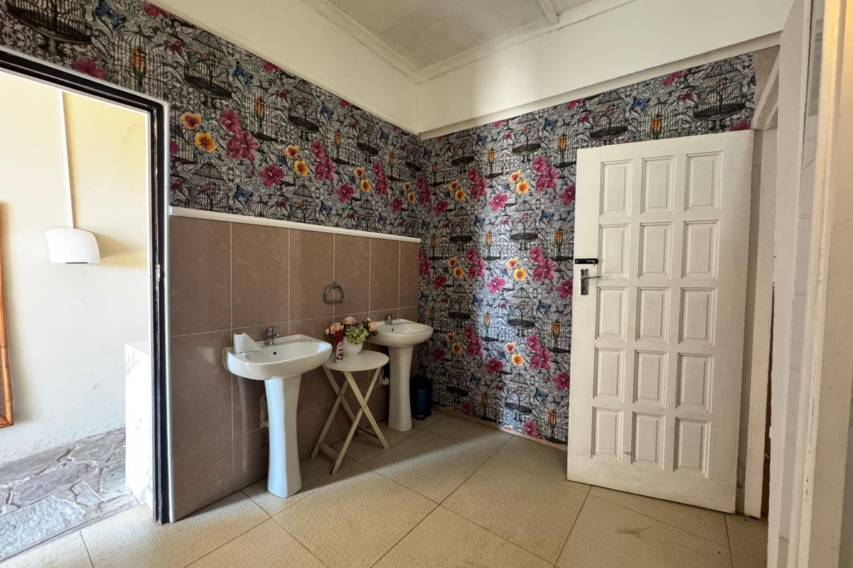 A functional bathroom area featuring dual pedestal wash basins, tiled splashbacks, and patterned wallpaper, requiring cosmetic updates to improve its overall condition.