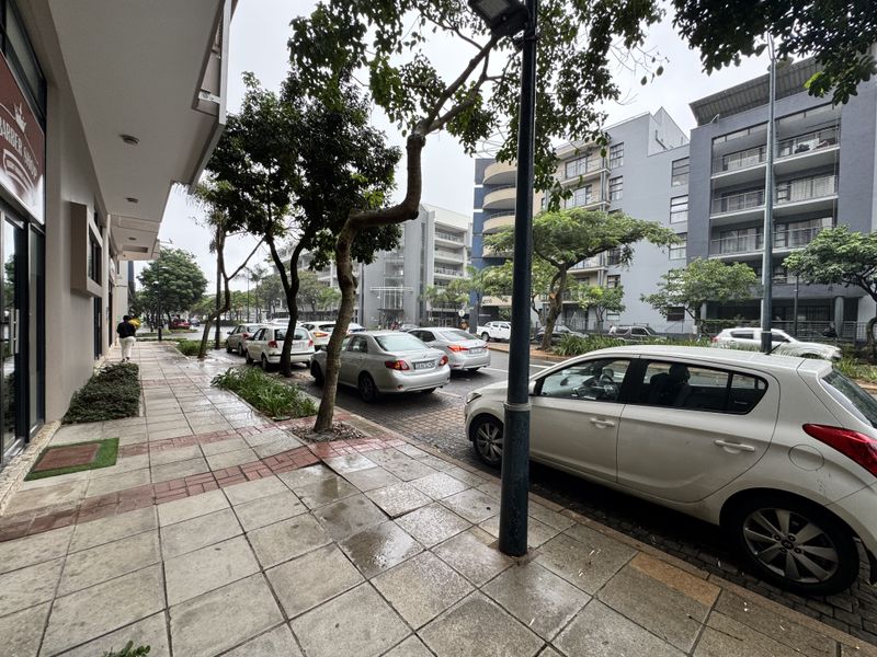 A street-level view of the retail property frontage, featuring a paved sidewalk, street lighting, and adjacent parking along the urban thoroughfare.