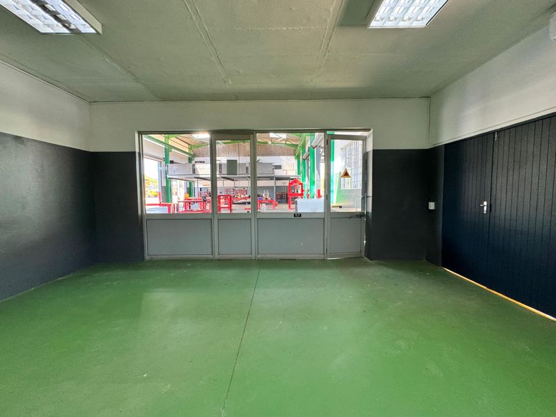 A clean, empty utility or office space featuring painted concrete flooring, fluorescent lighting, and a glass partition looking into the main industrial workshop area.
