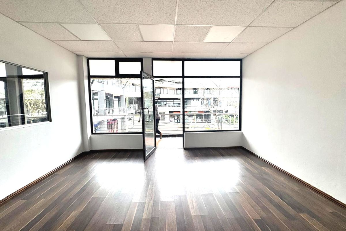 A bright, empty office space featuring modern laminate flooring, a suspended ceiling, and large windows providing natural light and access to a balcony.