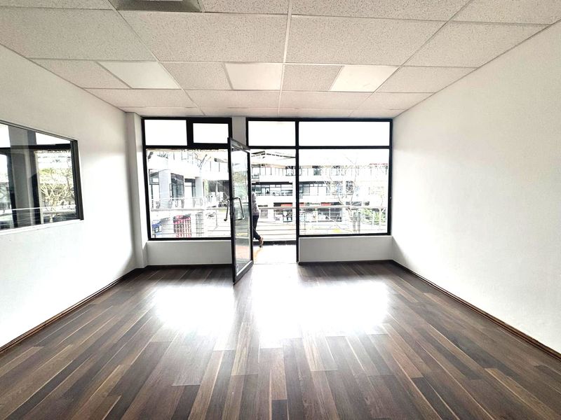 A bright, empty office space featuring modern laminate flooring, a suspended ceiling, and large windows providing natural light and access to a balcony.