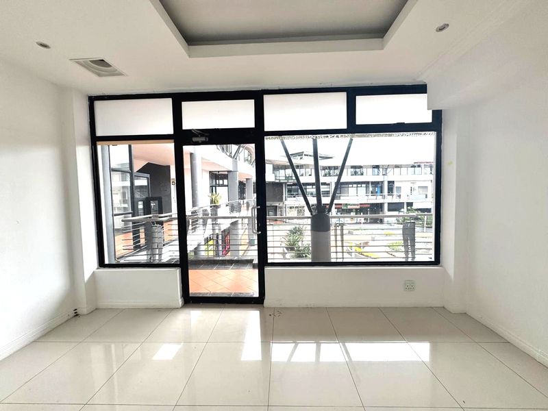 A bright, empty office space featuring large glass windows, a glass door, tiled flooring, and recessed ceiling lighting with a view of the surrounding complex.