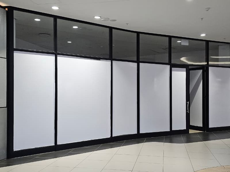 A vacant retail storefront featuring a modern glass facade with metal framing, recessed lighting, and tiled flooring in a shopping mall setting.