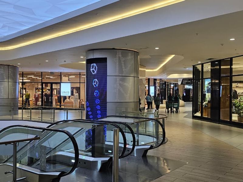 Modern retail shopping center interior featuring escalators, glass balustrades, and contemporary storefronts with integrated digital display columns and recessed lighting.